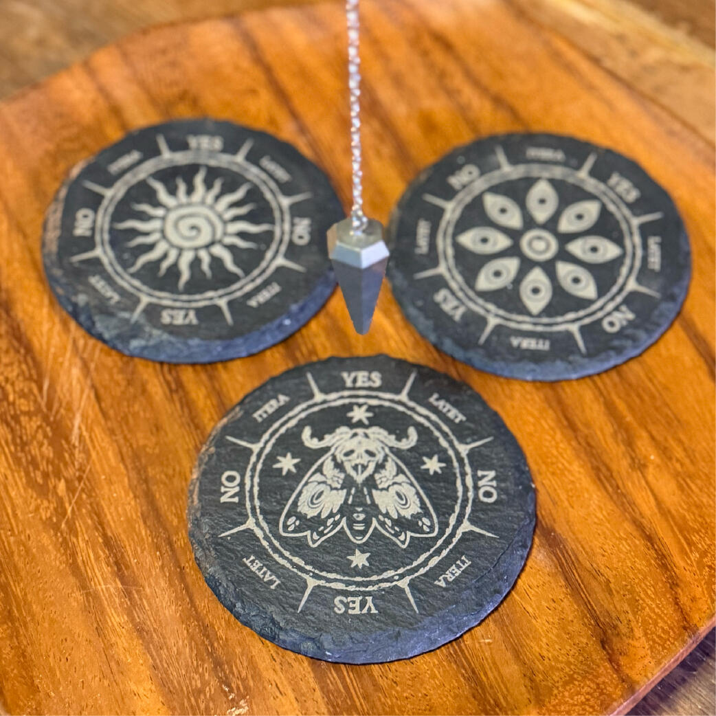 Markstones - Laid out to ask Engraved slate pendulum boards with sun, floral, and moth motifs, a pendulum at rest, Markstones by WMFrewolfe